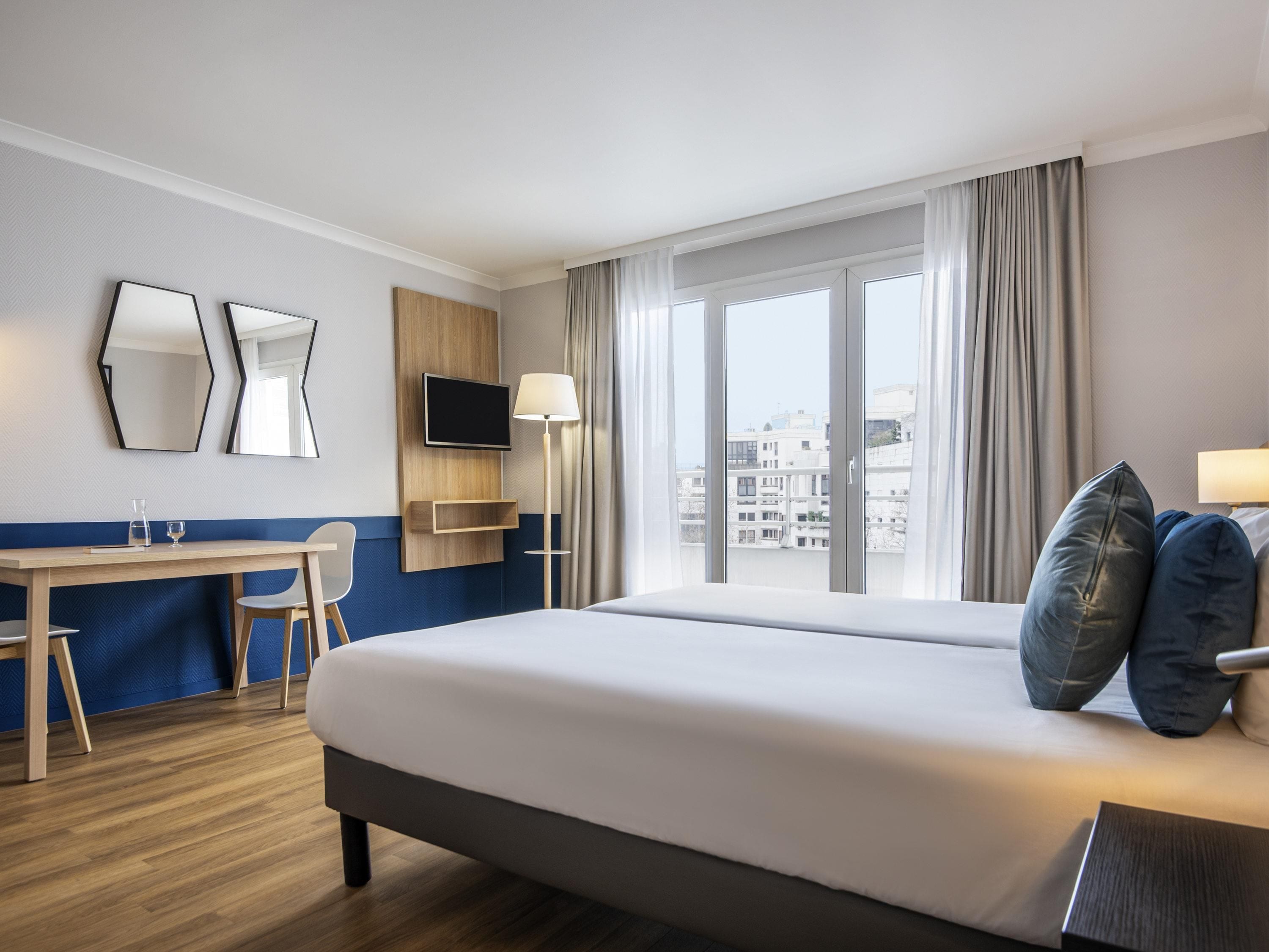 A hotel room with a bed, a desk, a chair, a TV, and a window with curtains.