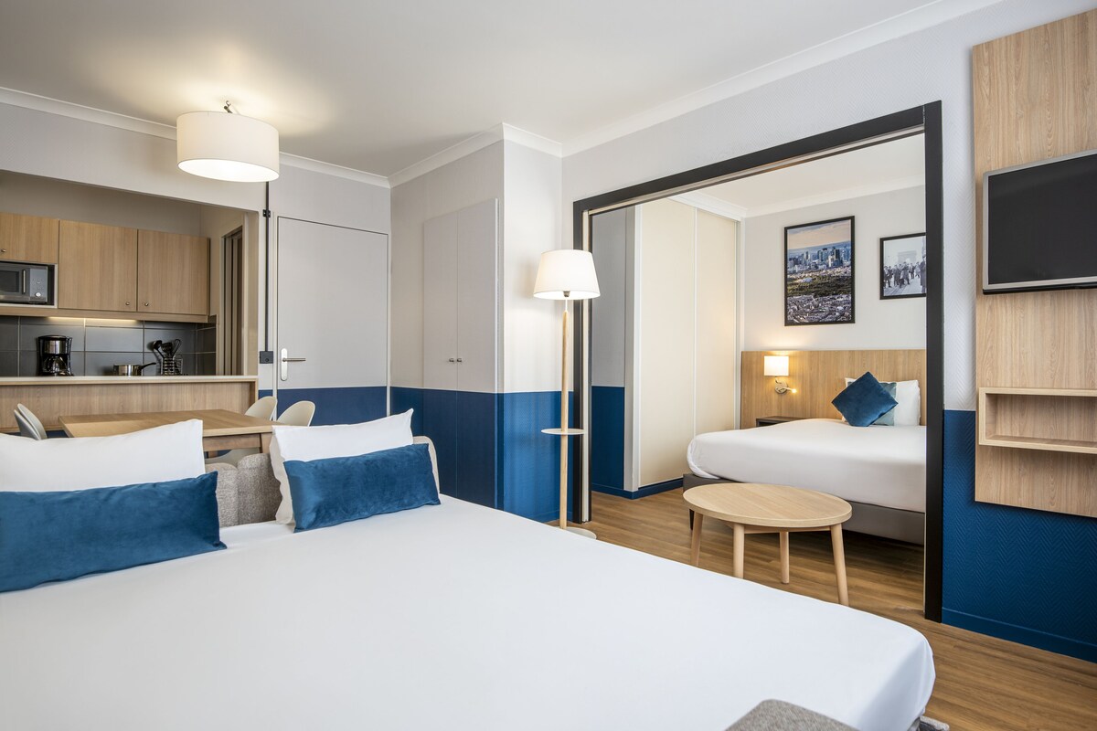 A hotel room with a large bed, a small table, a desk with a chair, a kitchenette, and a bathroom visible through an open door.
