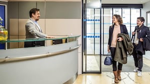 A reception desk with a glass partition, a tiled wall, and a door leading to a hallway with a patterned floor.