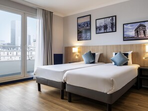 A hotel room with two beds, a large window with a view of buildings, and three framed pictures on the wall. - Aparthotel Adagio La Defense Kleber (Courbevoie)