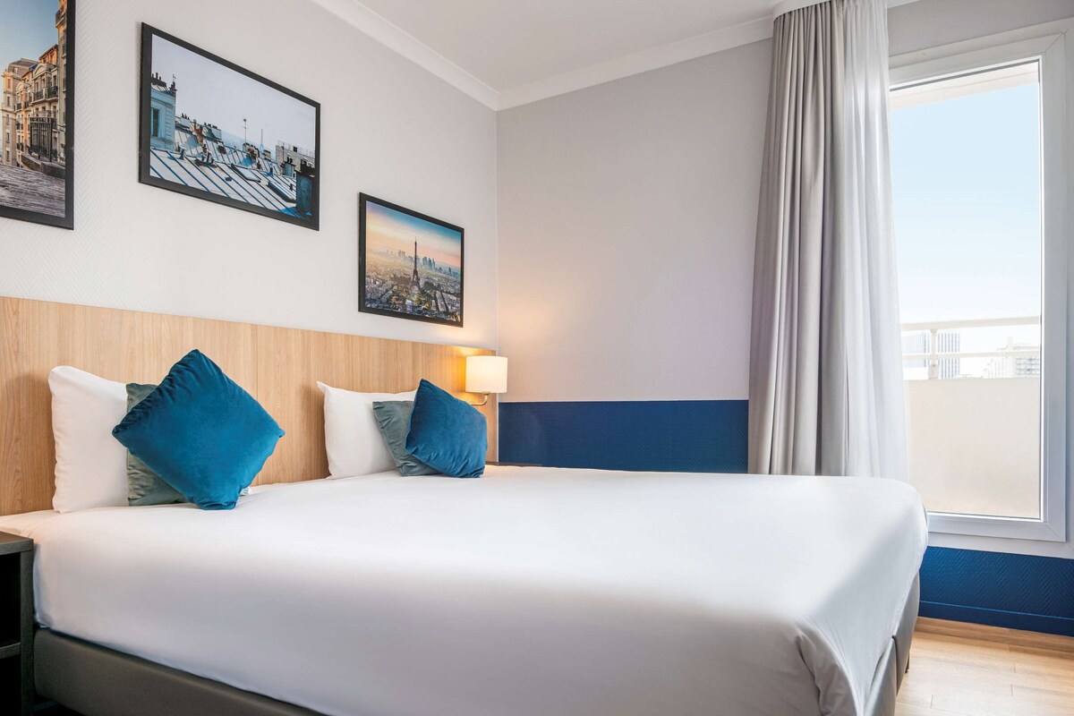 A hotel room with a large bed, two pillows, a nightstand, a wall-mounted lamp, and two framed pictures.