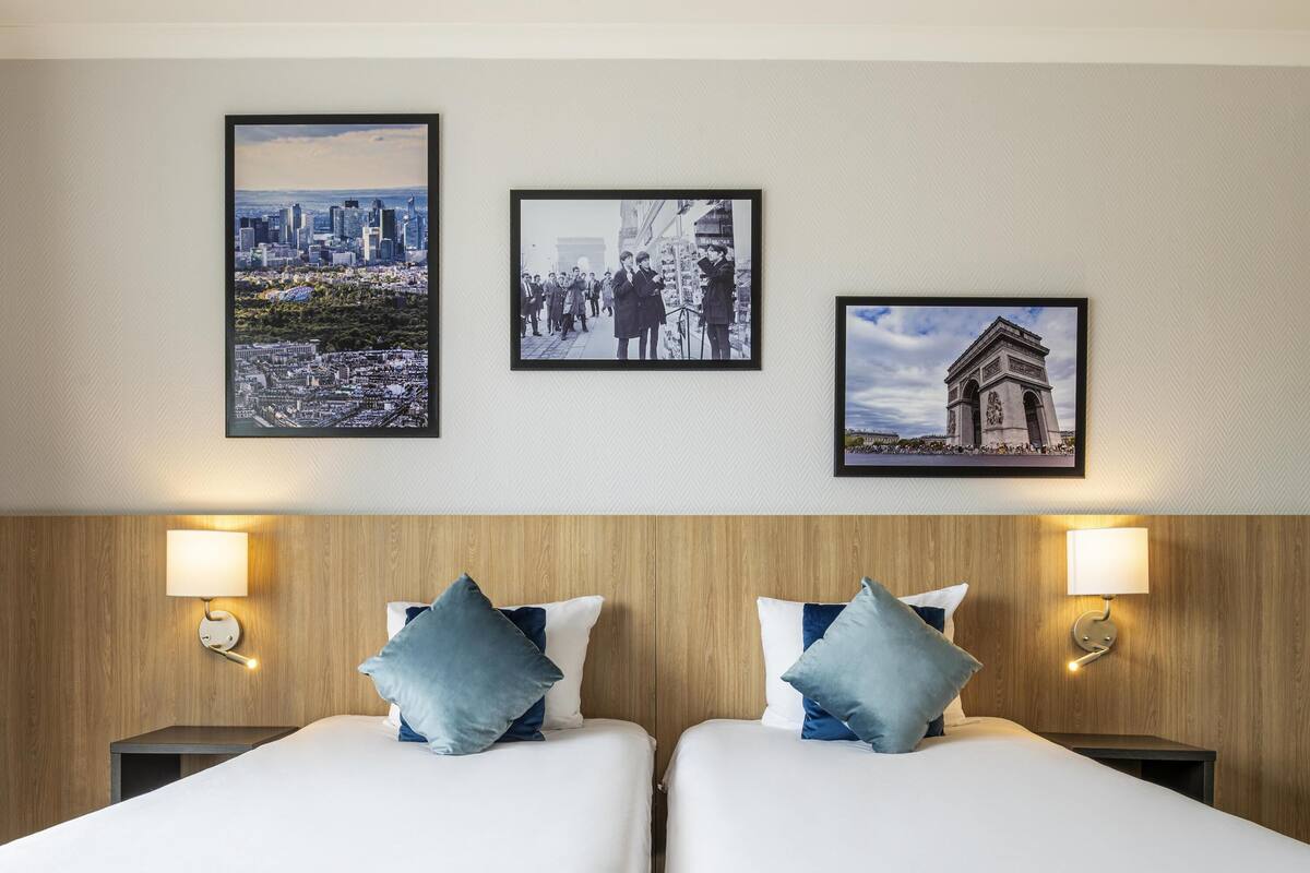 A hotel room with two beds, a wooden headboard, wall-mounted lamps, and three framed pictures.