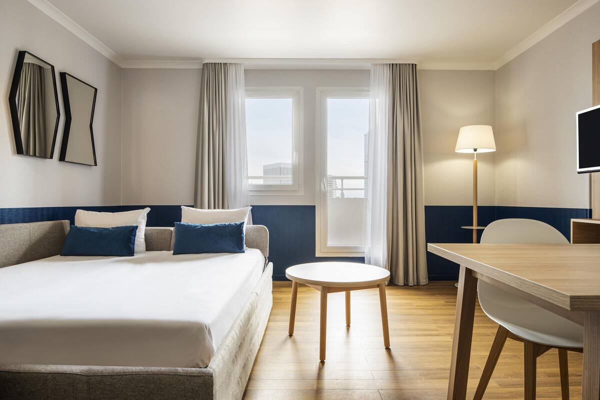 A hotel room with a bed, a round table, a chair, a desk, a lamp, and a TV.