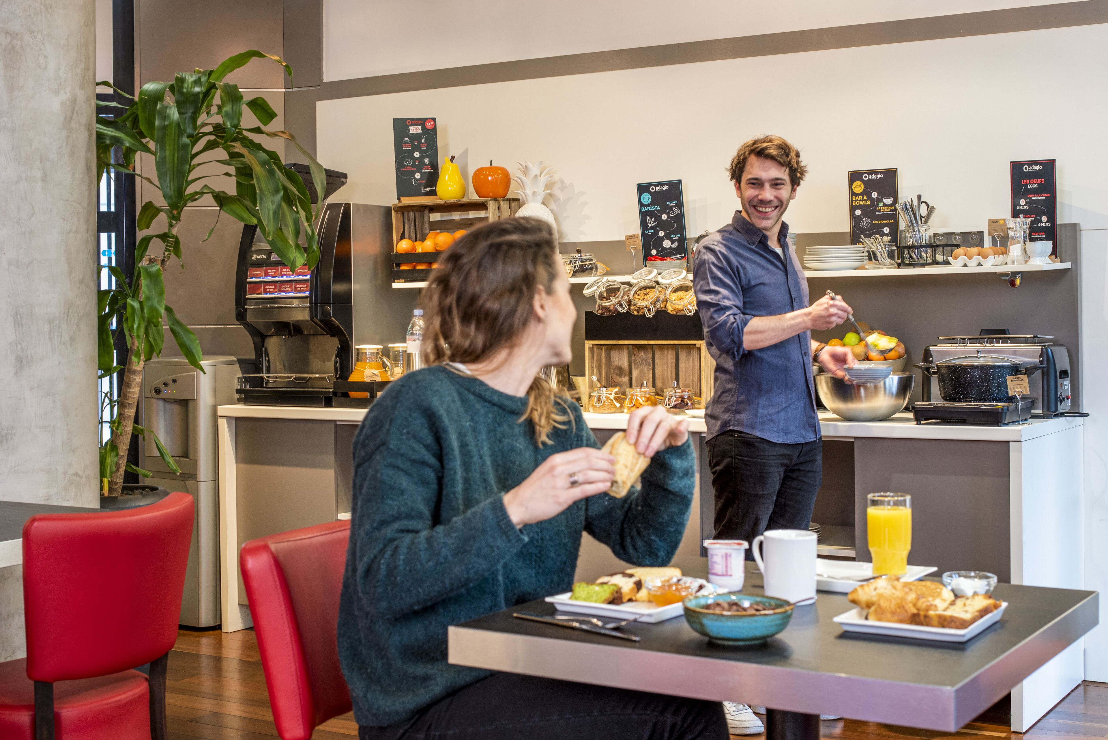 Un espace cuisine moderne comprenant une machine à café, un présentoir de fruits et un coin petit-déjeuner avec une table, des chaises et une personne qui sert les aliments.