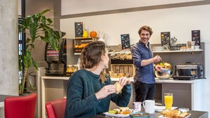 A modern kitchen area with a coffee machine, a fruit display, and a breakfast setting with a table, chairs, and a person serving food. - Aparthotel Adagio La Defense Kleber (Courbevoie)