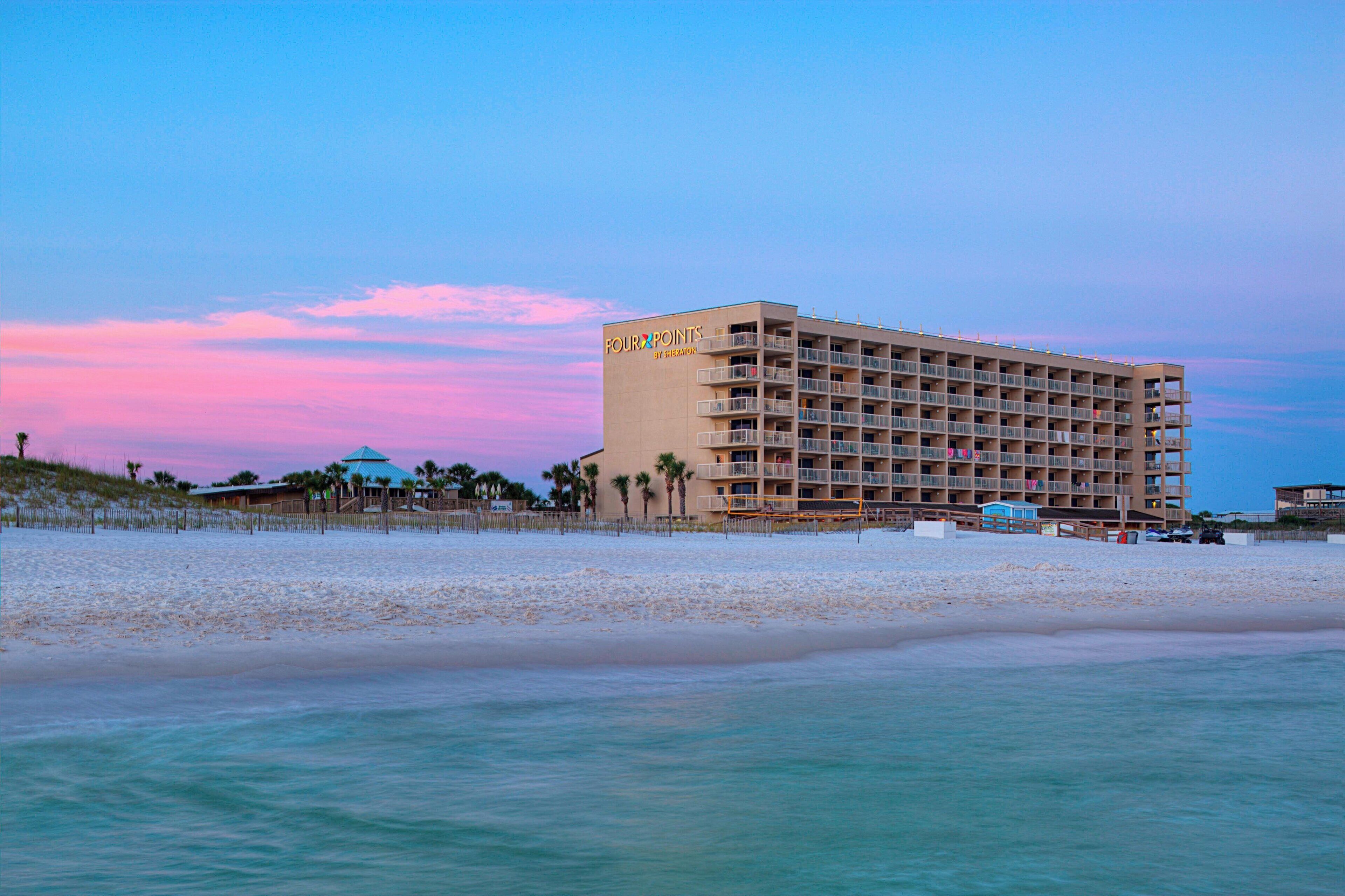 Un hôtel situé en bord de mer, doté de nombreux balcons, d’une plage de sable fin et offrant une vue sur un ciel dégagé.