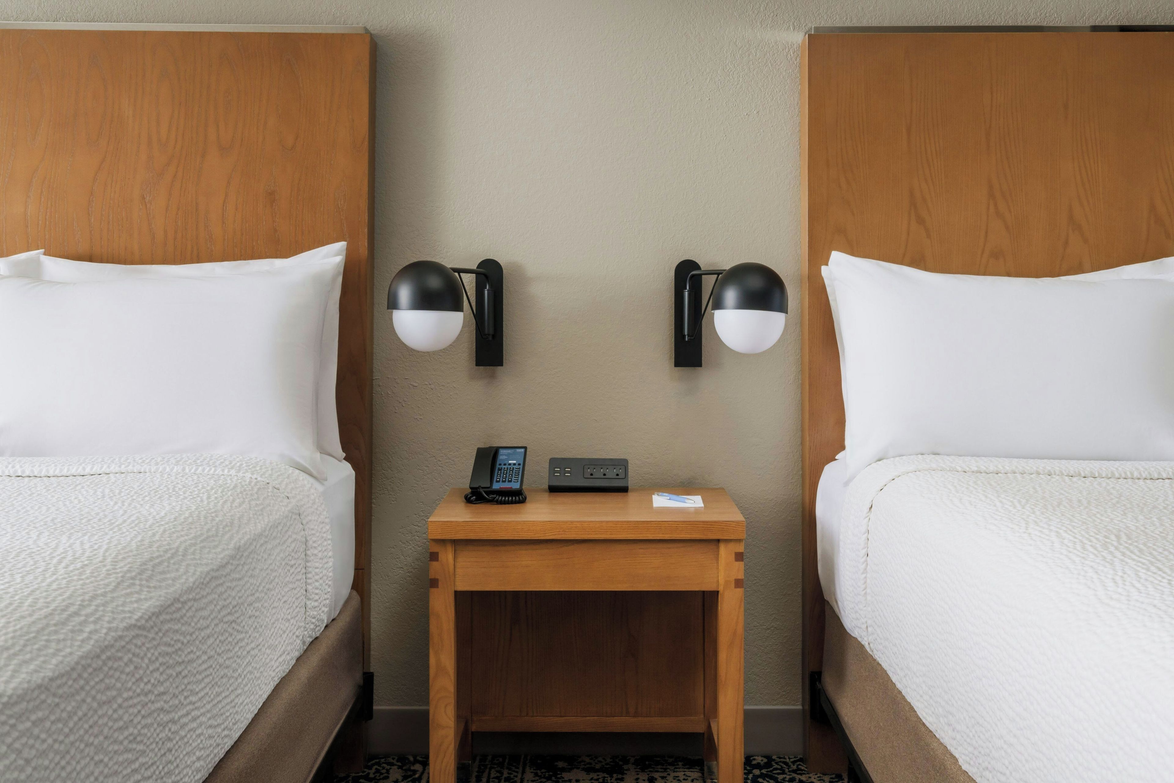 Habitación de hotel con dos camas, una mesita de noche de madera, un teléfono y dos lámparas fijadas a la pared.