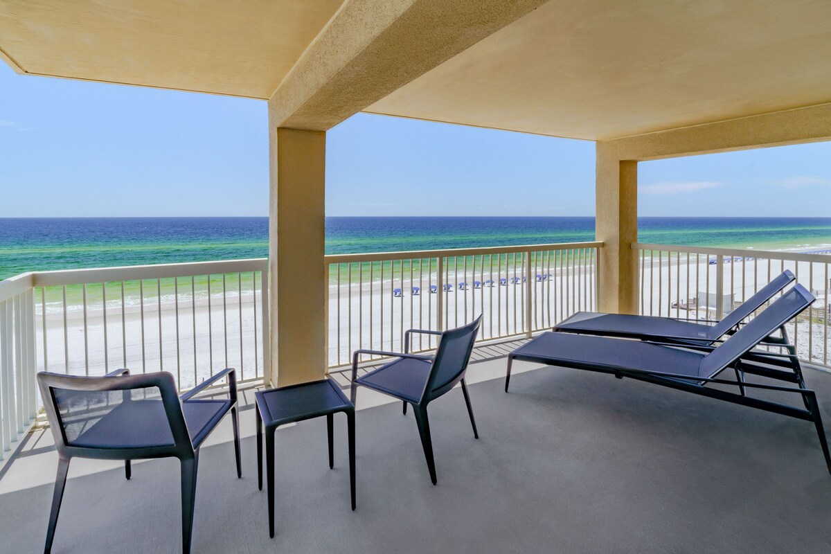 A covered outdoor area with a view of the beach and ocean. There are two lounge chairs and two tables.