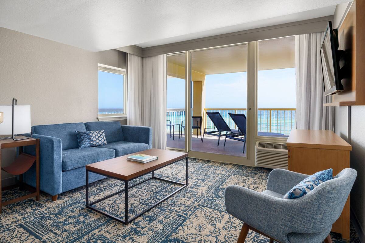 Een hotelkamer met een bank, fauteuil en een salontafel. Er is een balkon met uitzicht op de oceaan.