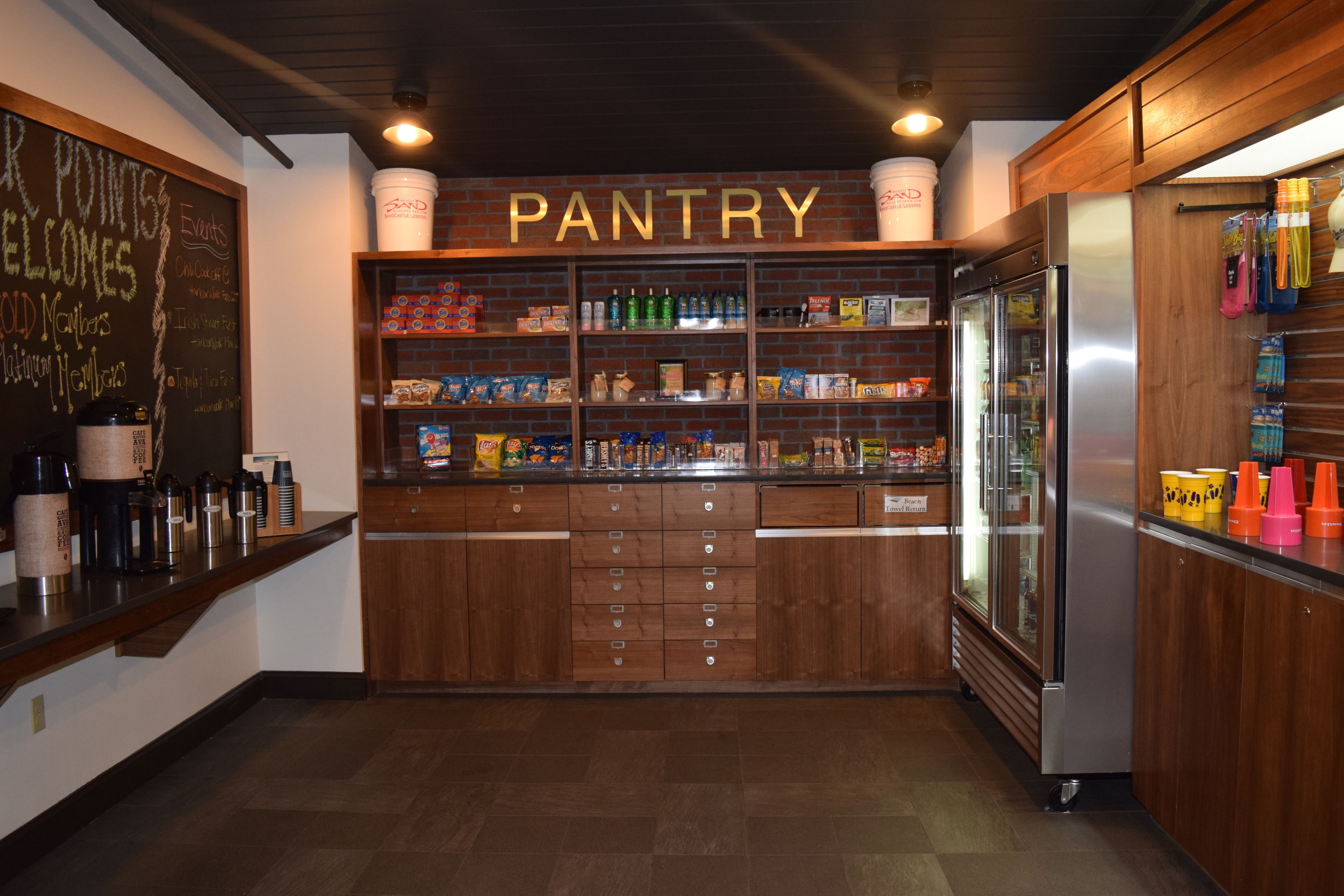 A pantry with shelves stocked with snacks and beverages, a chalkboard with event information, and a refrigerator.
