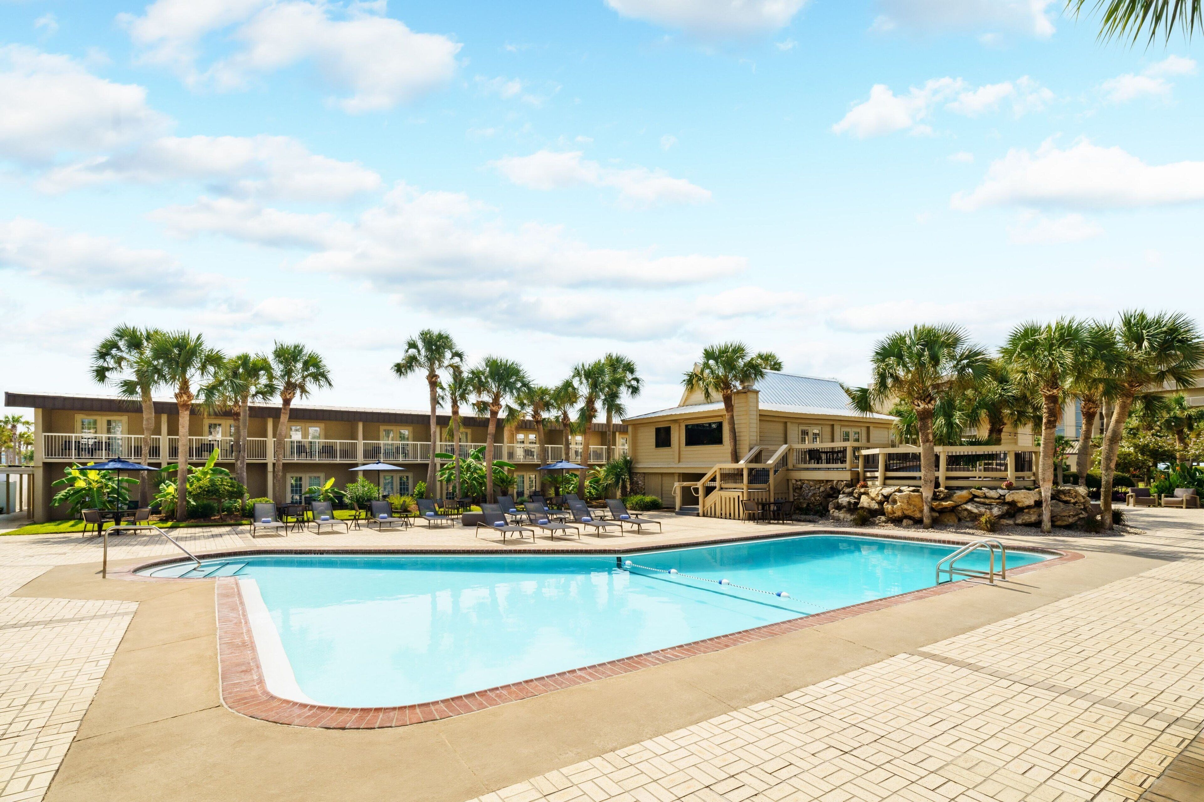 A swimming pool with lounge chairs and umbrellas, surrounded by palm trees and a building with balconies.