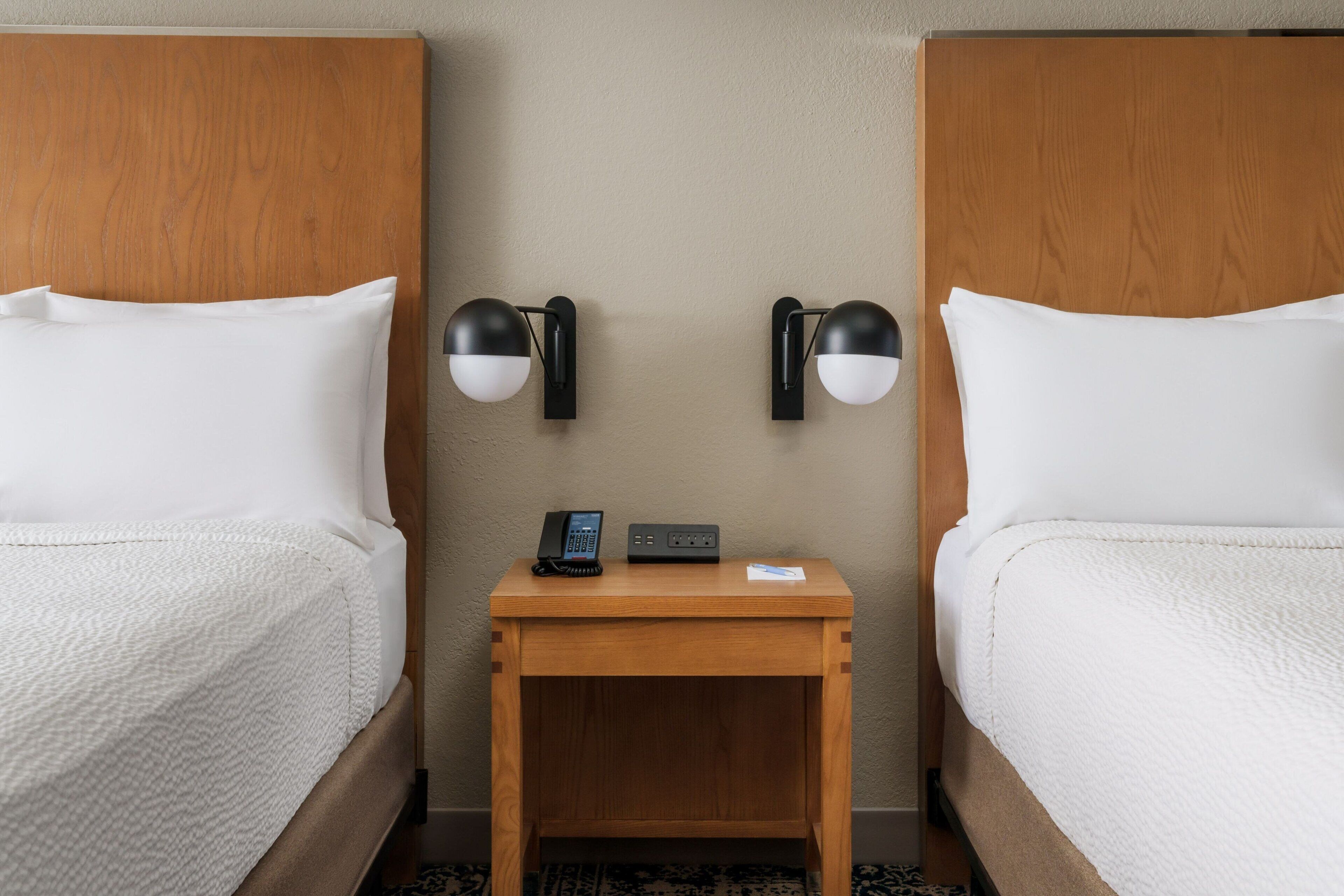 A hotel room with two beds, a wooden nightstand with a phone and a small table, and wall-mounted lamps.