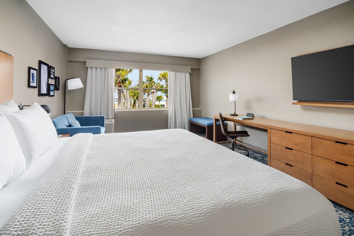A hotel room with a large bed, a desk, a chair, a TV, and a window with curtains.