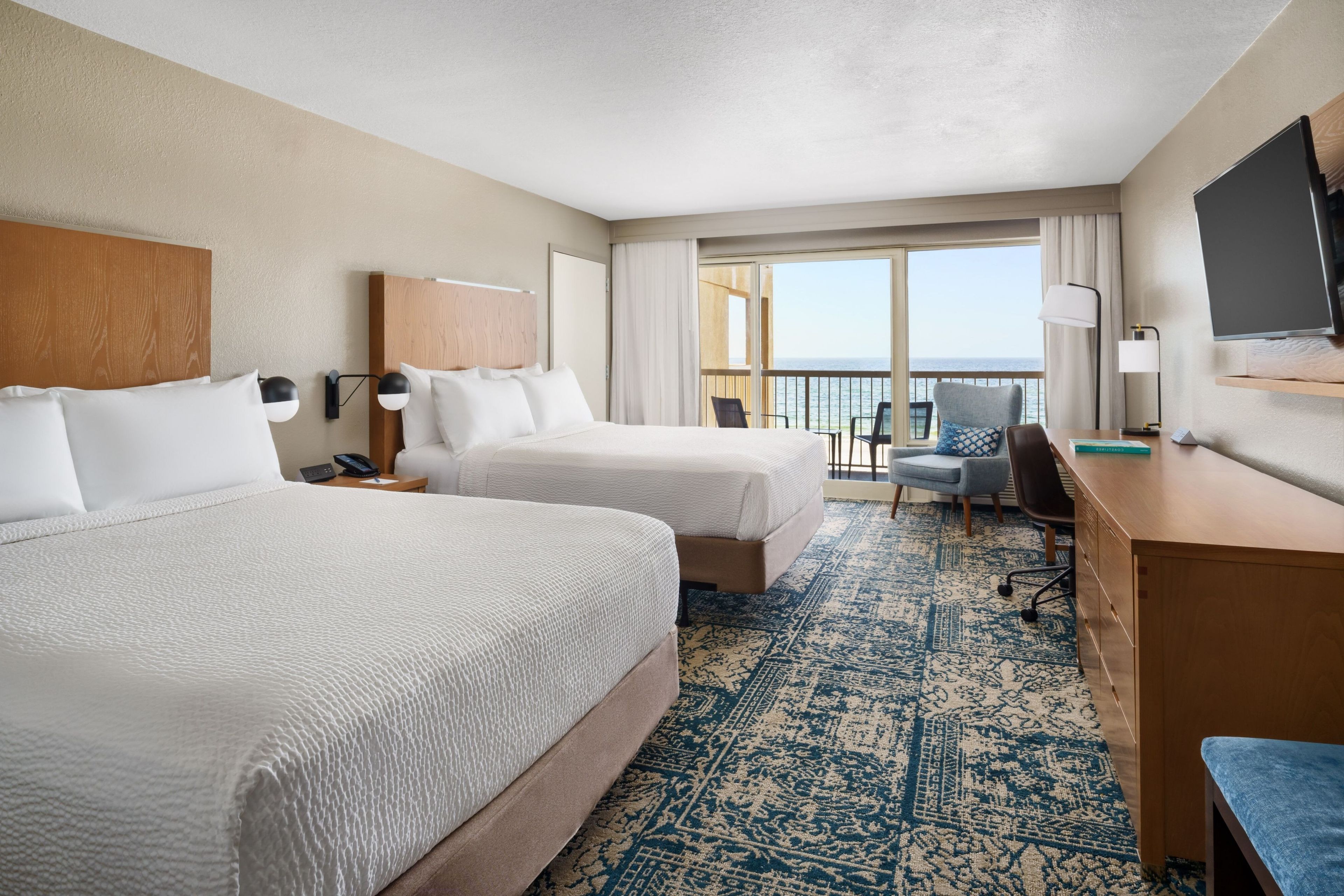 Habitación de hotel con dos camas, un escritorio, una silla, un televisor y un balcón con vistas al mar.