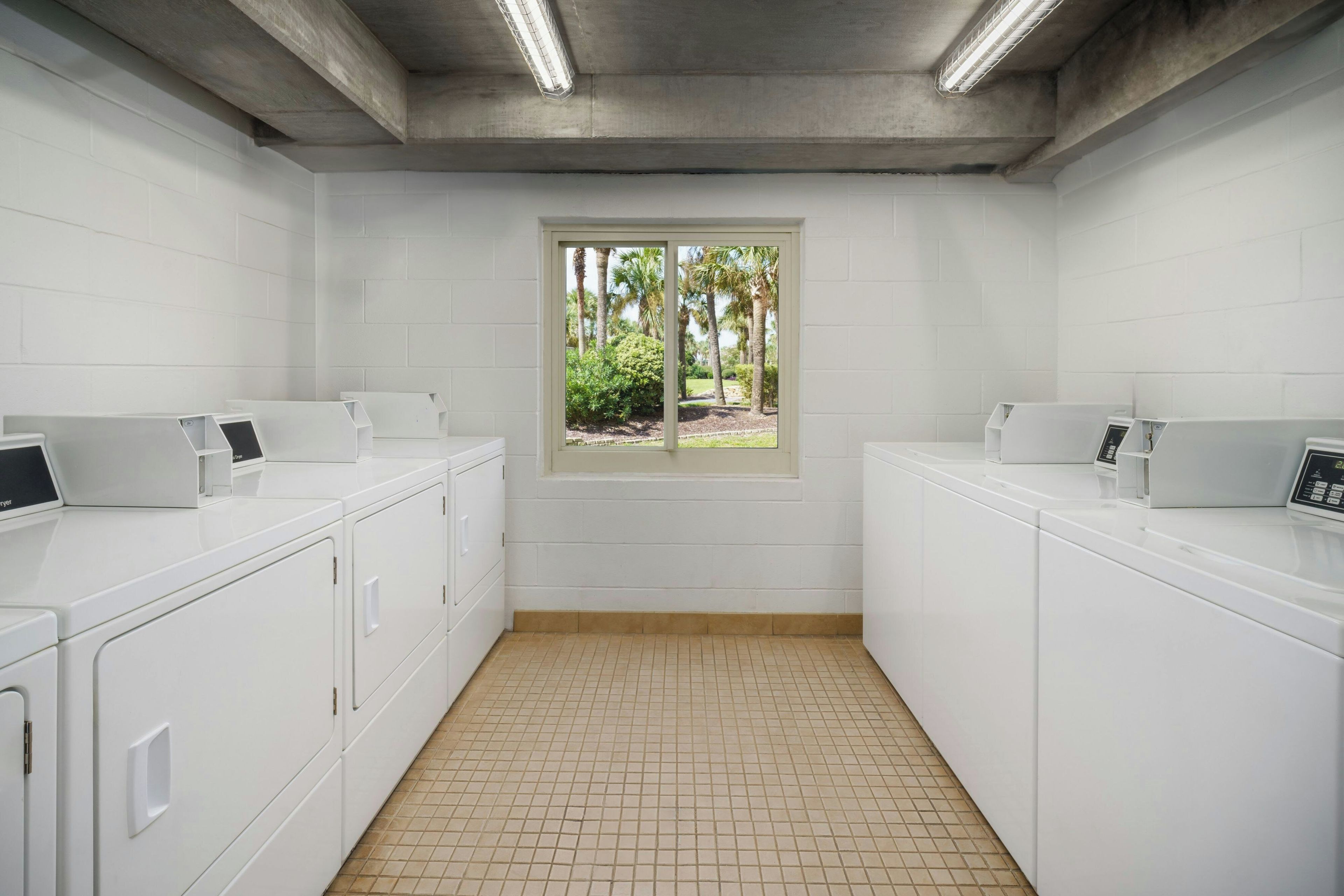A laundry room with two washing machines, a window with a view of palm trees, and a tiled floor.