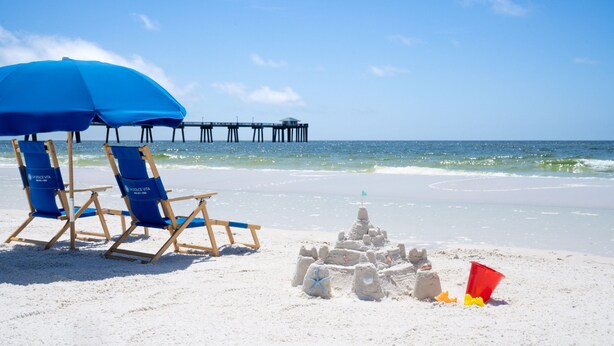 Escena de playa con un castillo de arena, sillas de playa azules y un muelle al fondo.