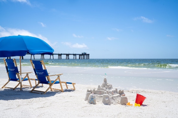 A beach scene with a sandcastle, blue beach chairs, and a pier in the background. - Four Points by Sheraton Destin-Fort Walton Beach (Fort Walton Beach)