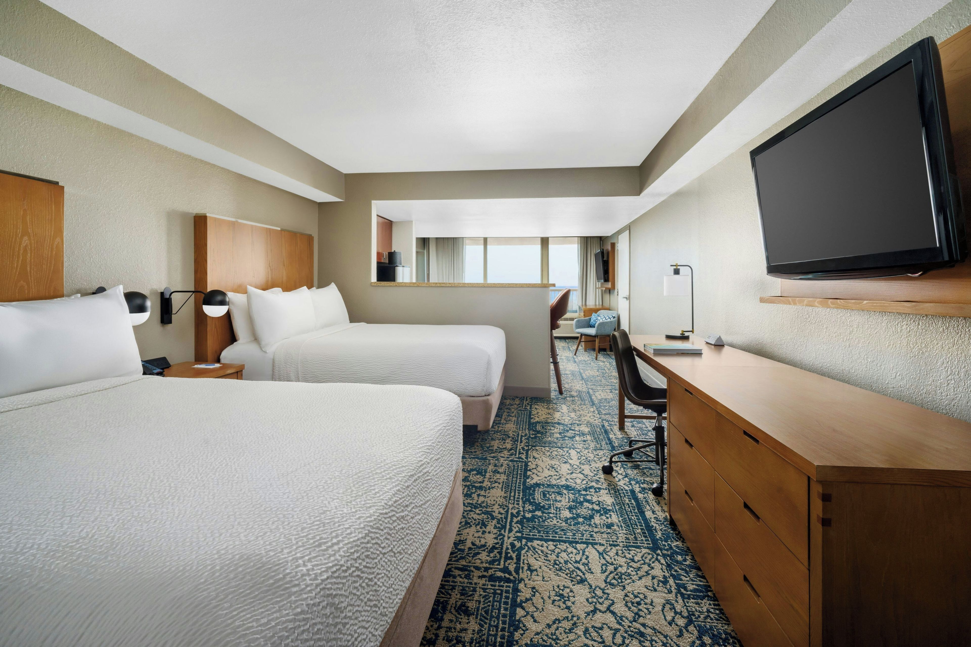 A hotel room with two beds, a desk, a television, and a view of the outside.