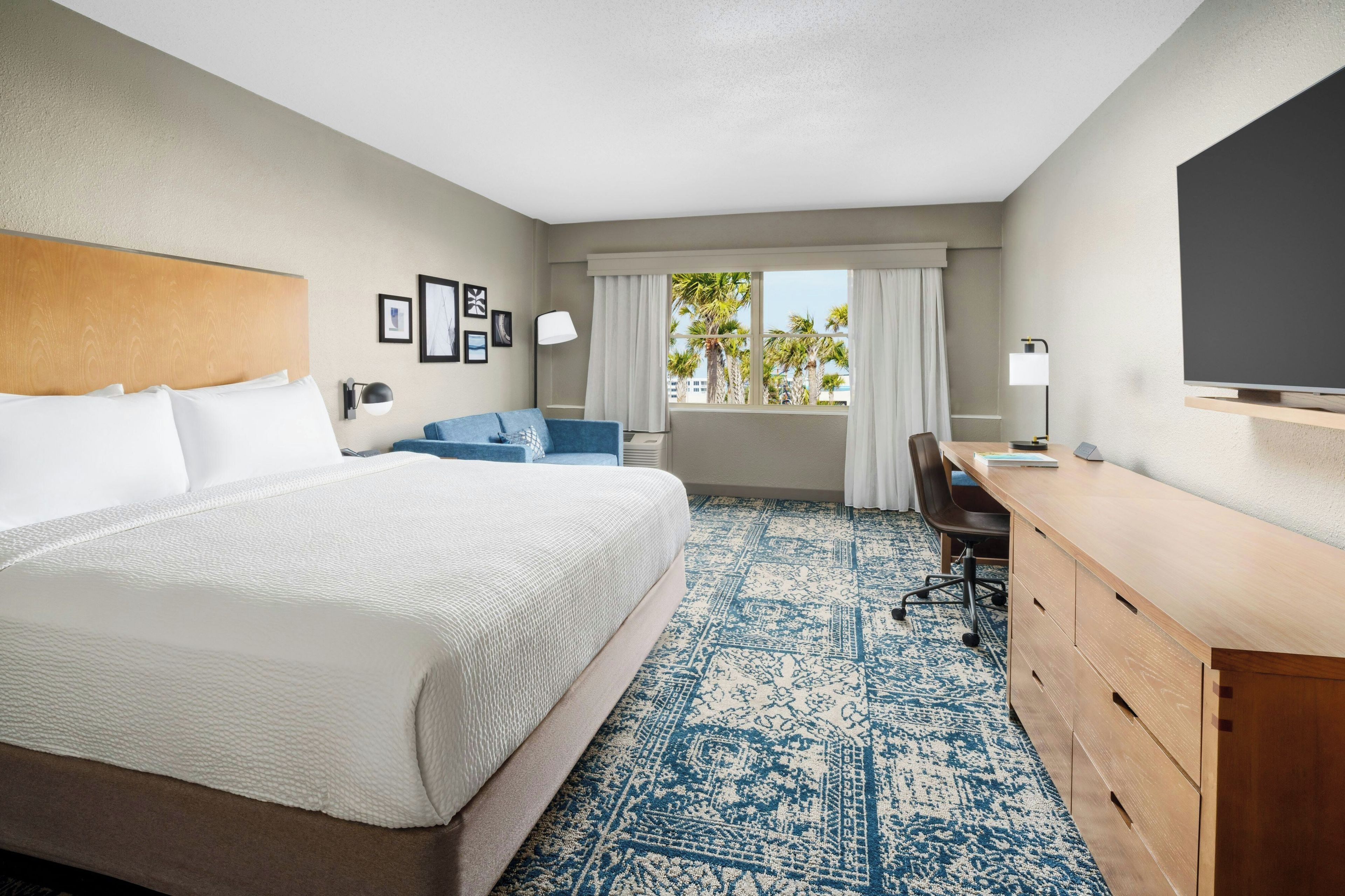 A hotel room with a large bed, a desk, a chair, a TV, and a window with a view of palm trees.