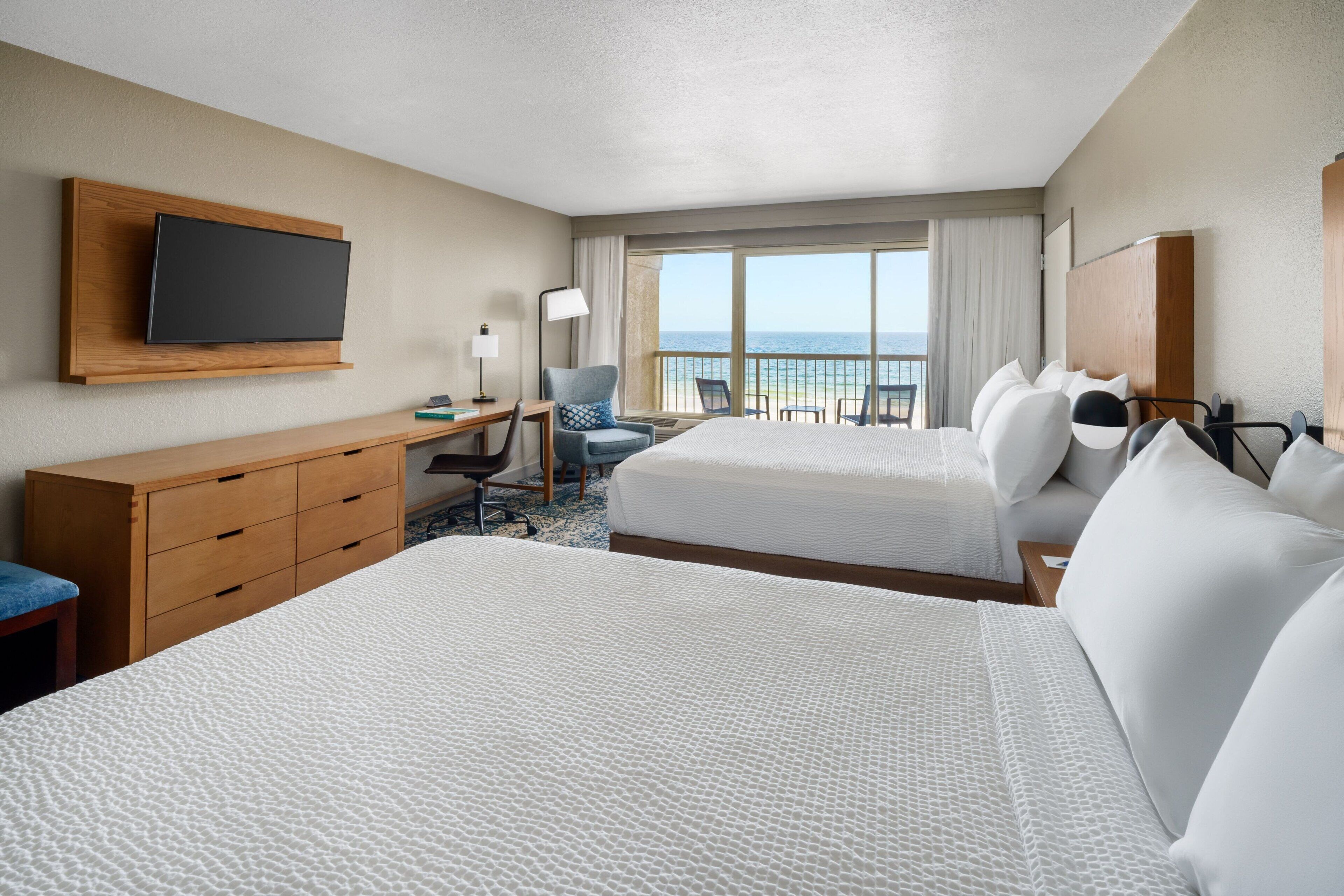 A hotel room with two beds, a desk, a chair, a TV, and a view of the ocean.