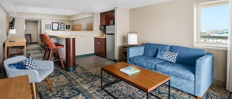 A modern living room with a blue sofa, a wooden coffee table, a patterned rug, and a view of the city through a window.