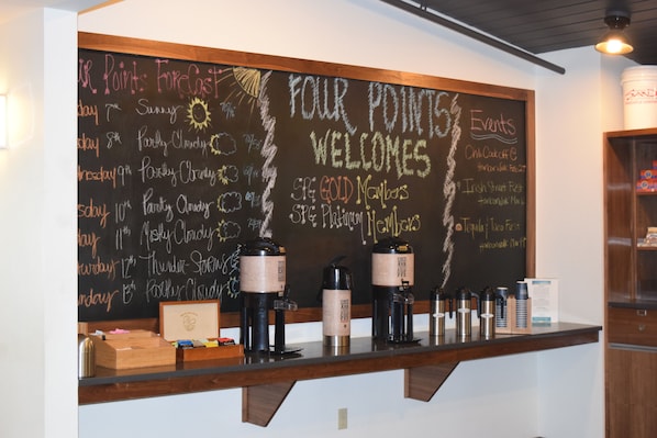 A chalkboard with weather forecasts and event listings, a coffee station with thermos cups, and a wooden counter.