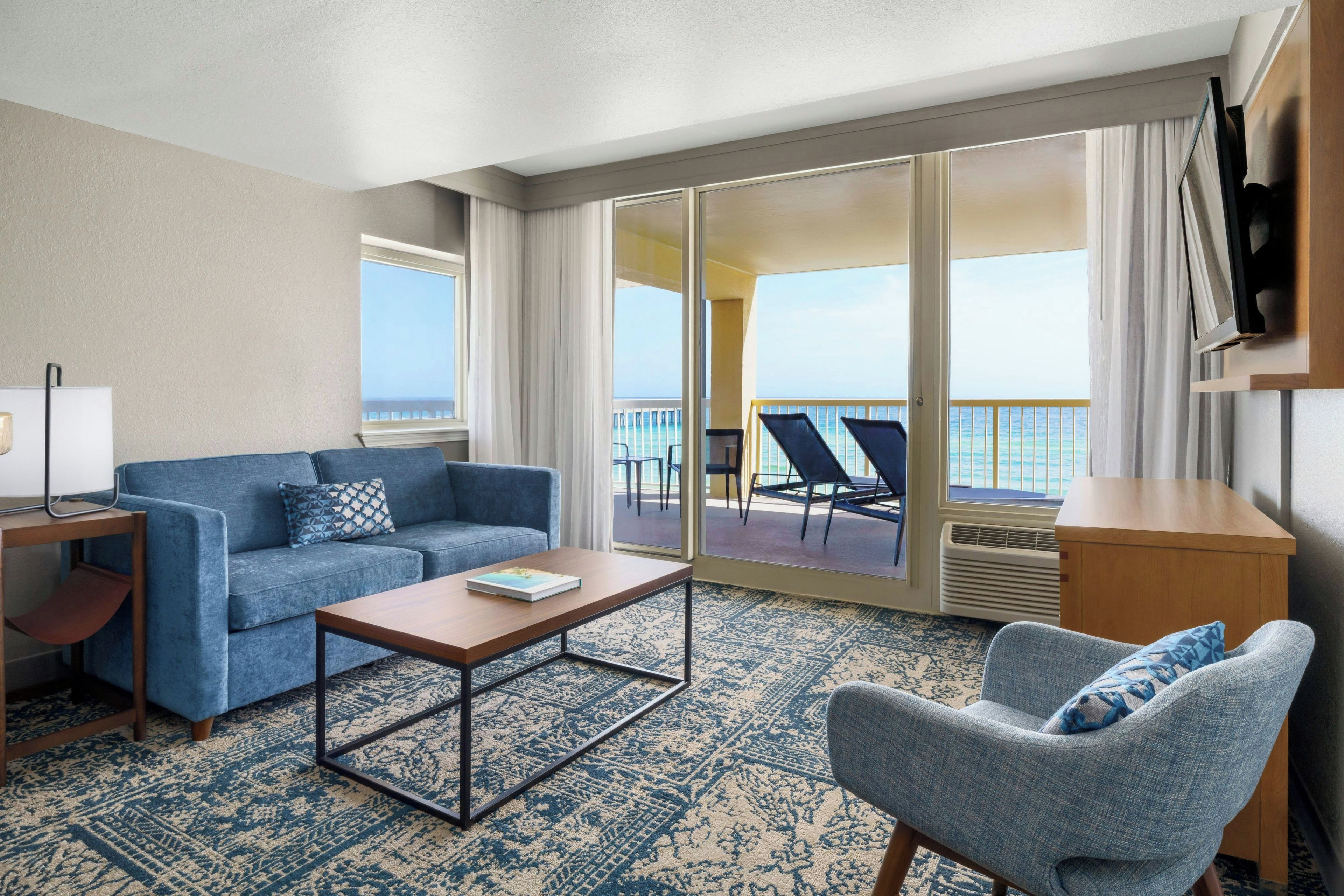 Habitación de hotel con sofá, sillón, mesa de centro y vista al océano a través de amplias puertas correderas.
