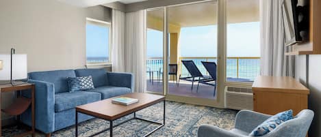Habitación de hotel con sofá, sillón, mesa de centro y vista al océano a través de amplias puertas correderas.