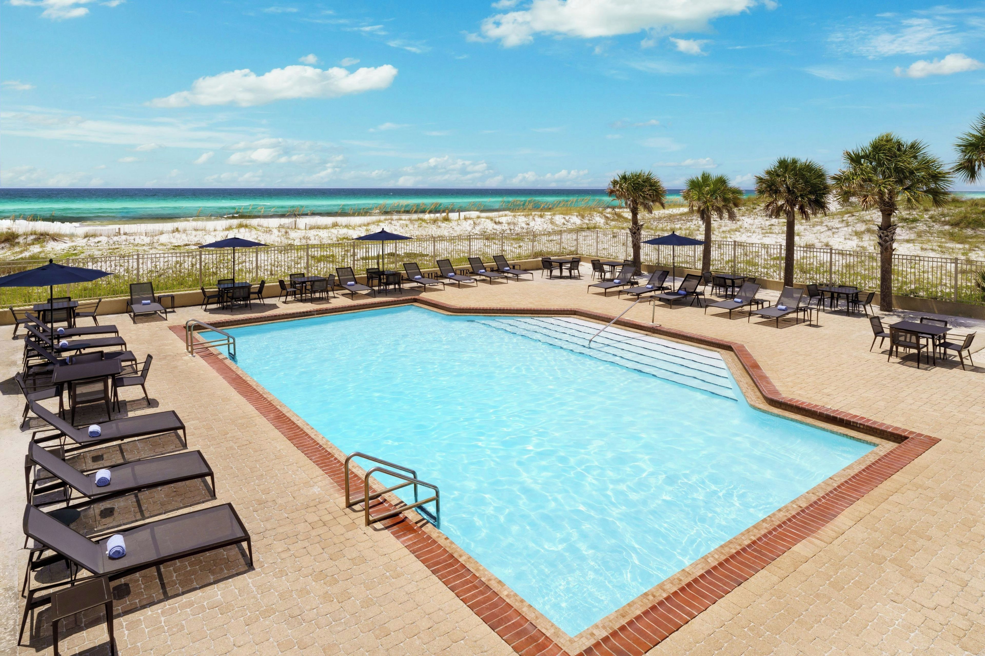 A rectangular swimming pool with a brick border, surrounded by lounge chairs and umbrellas, with a sandy beach and ocean view in the background.