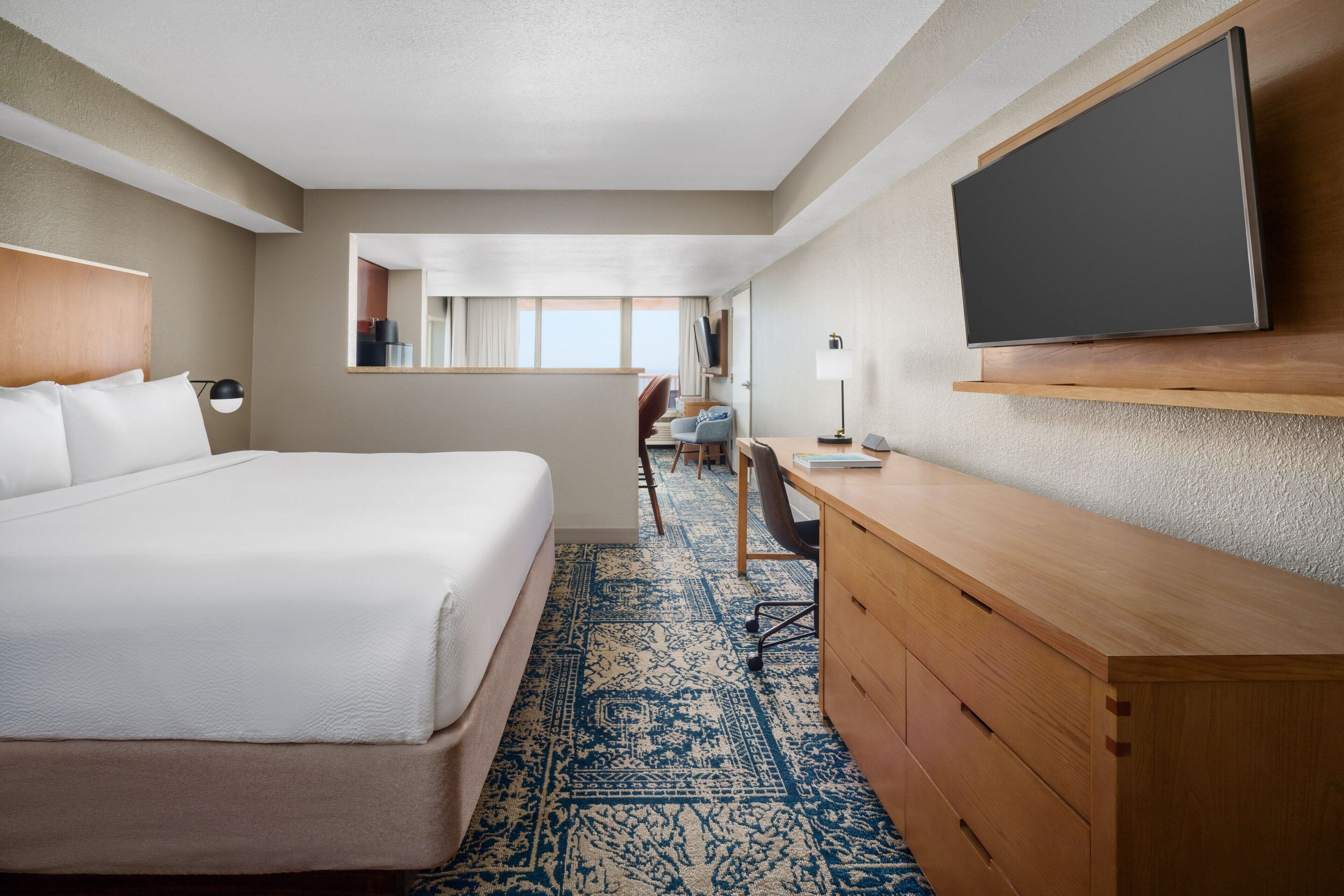 A hotel room with a large bed, a desk, a chair, a flat-screen TV mounted on the wall, and a patterned carpet.