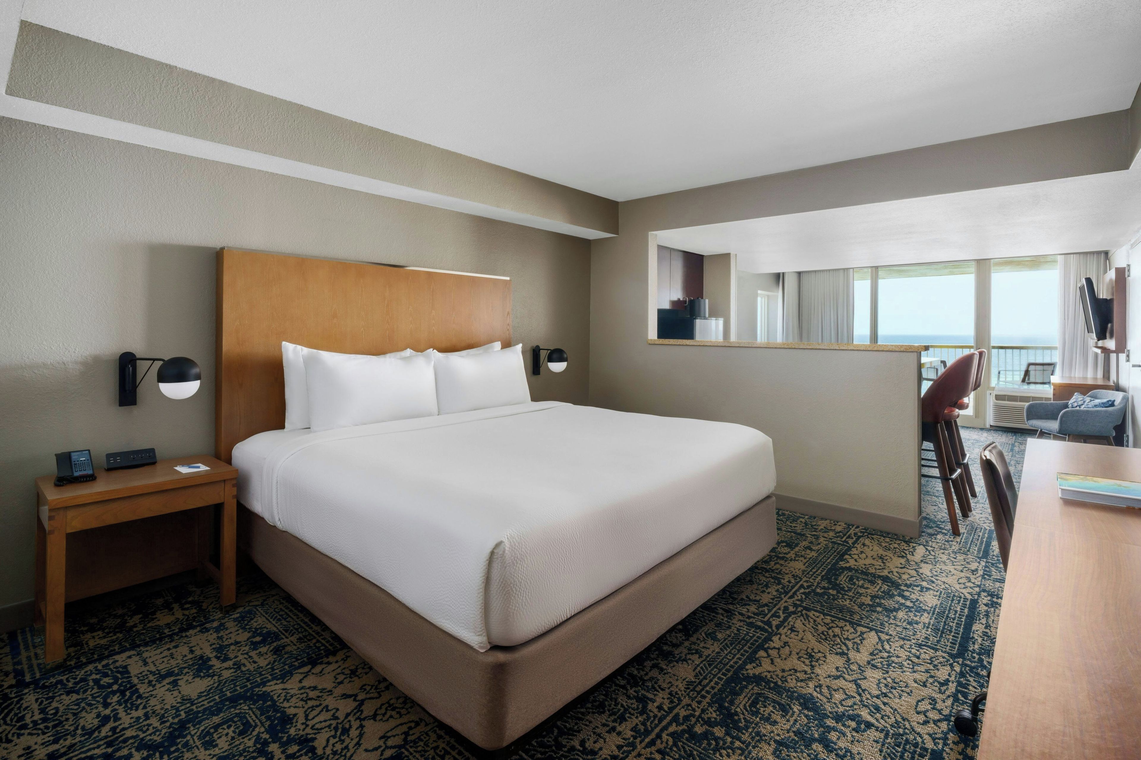 A hotel room with a large bed, a desk, a chair, a mirror, and a view of the sea.
