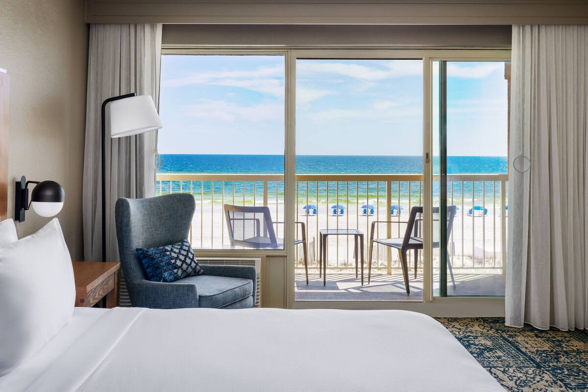 Een hotelkamer met een groot bed, een stoel, een tafel en uitzicht op het strand en de oceaan door een schuifdeur.