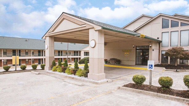 Entrada de hotel con zona de estacionamiento techada, señal de estacionamiento para discapacitados y un edificio con varias ventanas y un balcón.