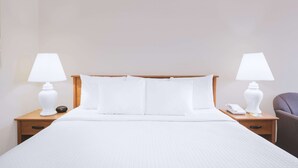 Una cama bien hecha con sábanas blancas, dos mesitas de noche con lámparas, una silla y una mesita con un teléfono y un libro.