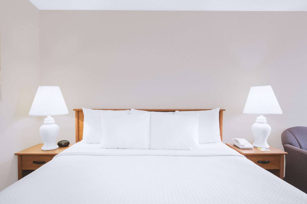 A neatly made bed with white linens, two bedside tables with lamps, a chair, and a nightstand with a phone and a book.