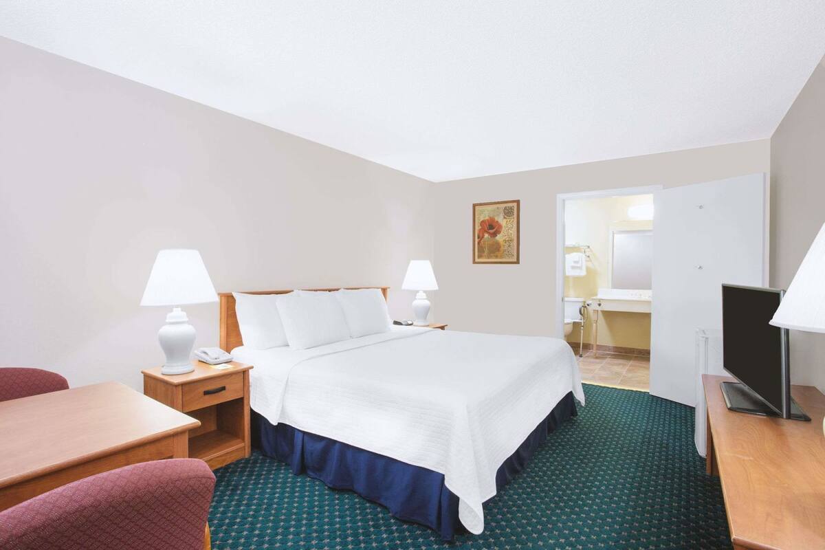 A hotel room with a bed, bedside tables, a desk, a chair, a TV, and a bathroom visible through an open door.