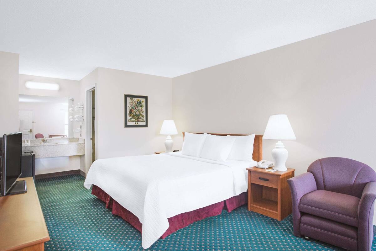 A hotel room with a bed, two bedside lamps, a chair, a desk with a computer, and a bathroom visible through an open door.
