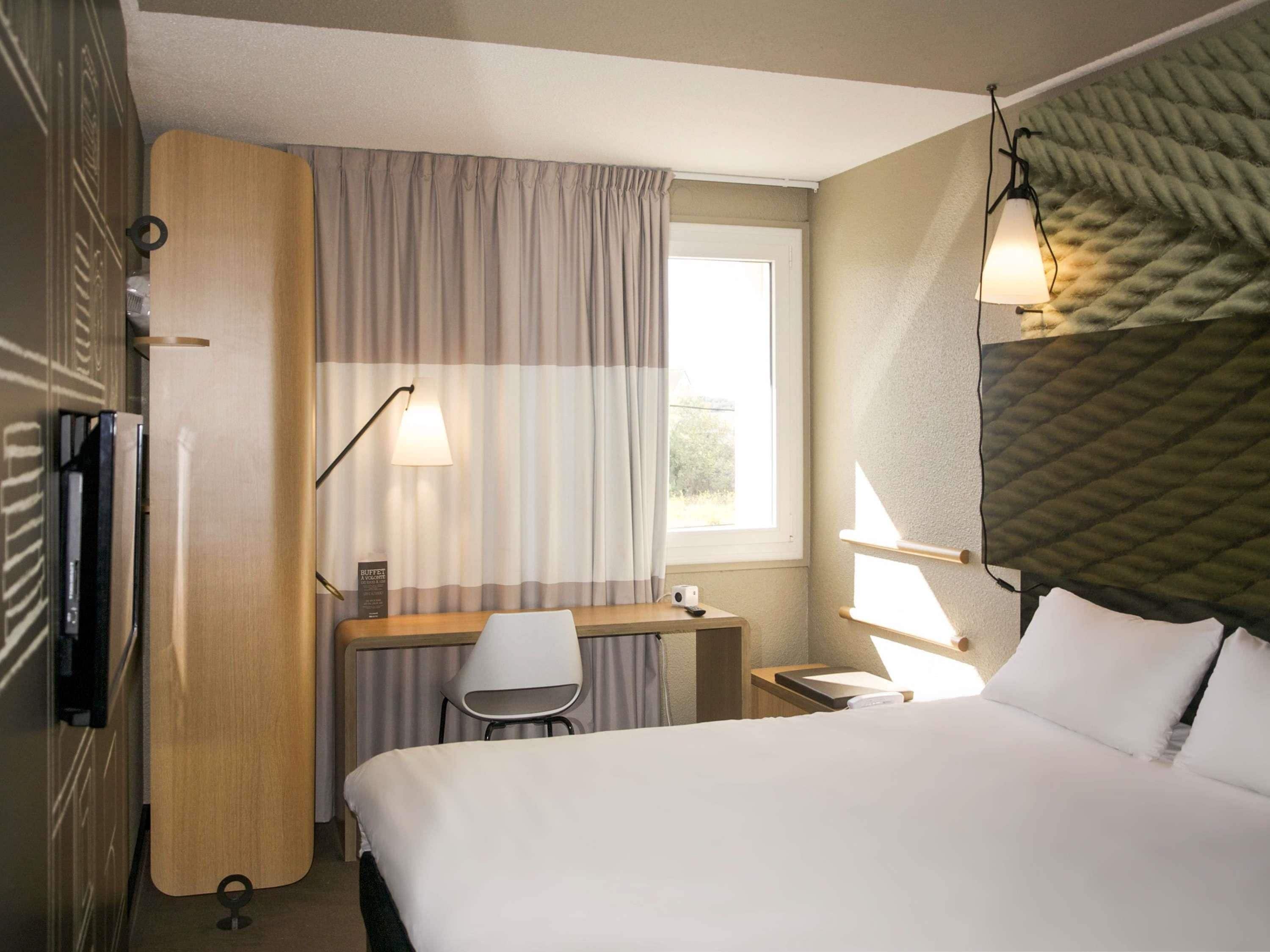 Una habitación de hotel con cama, escritorio, silla, lámpara y ventana con cortinas.