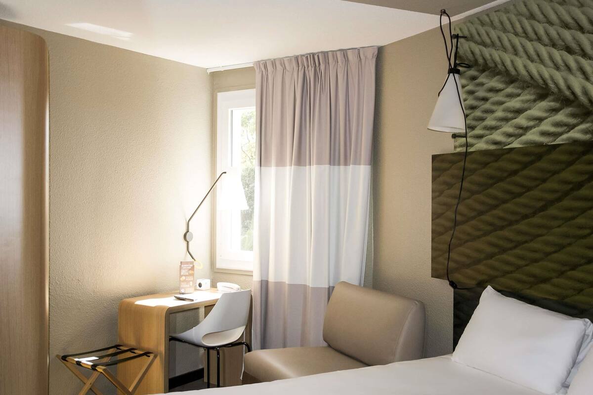 Une chambre d’hôtel avec un lit, un bureau avec une chaise, une lampe et une fenêtre avec des rideaux.