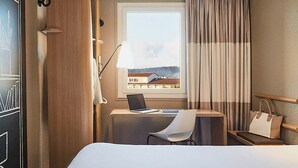 A hotel room with a bed, a desk with a laptop, a chair, a window with curtains, and a handbag on a side table. - ibis Toulon La Seyne Sur Mer (La Seyne-sur-Mer)