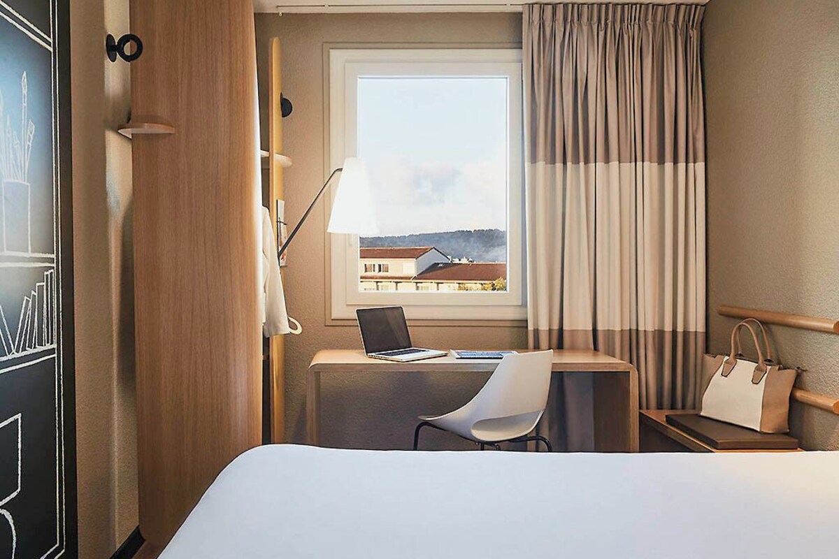 A hotel room with a bed, a desk with a laptop, a chair, a window with curtains, and a handbag on a side table.