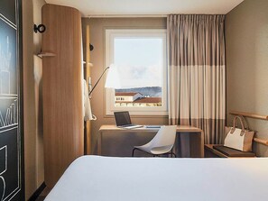 Hotelkamer met een bed, een bureau met een laptop, een stoel, een raam met gordijnen en een handtas op een bijzettafeltje.