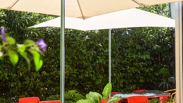 An outdoor seating area with tables and chairs, a large umbrella, and dense foliage in the background.