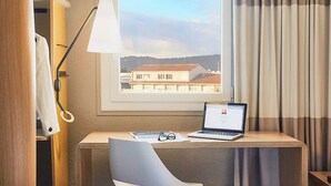 A hotel room with a desk, laptop, and chair, a window with curtains, and a view of buildings. - ibis Toulon La Seyne Sur Mer (La Seyne-sur-Mer)