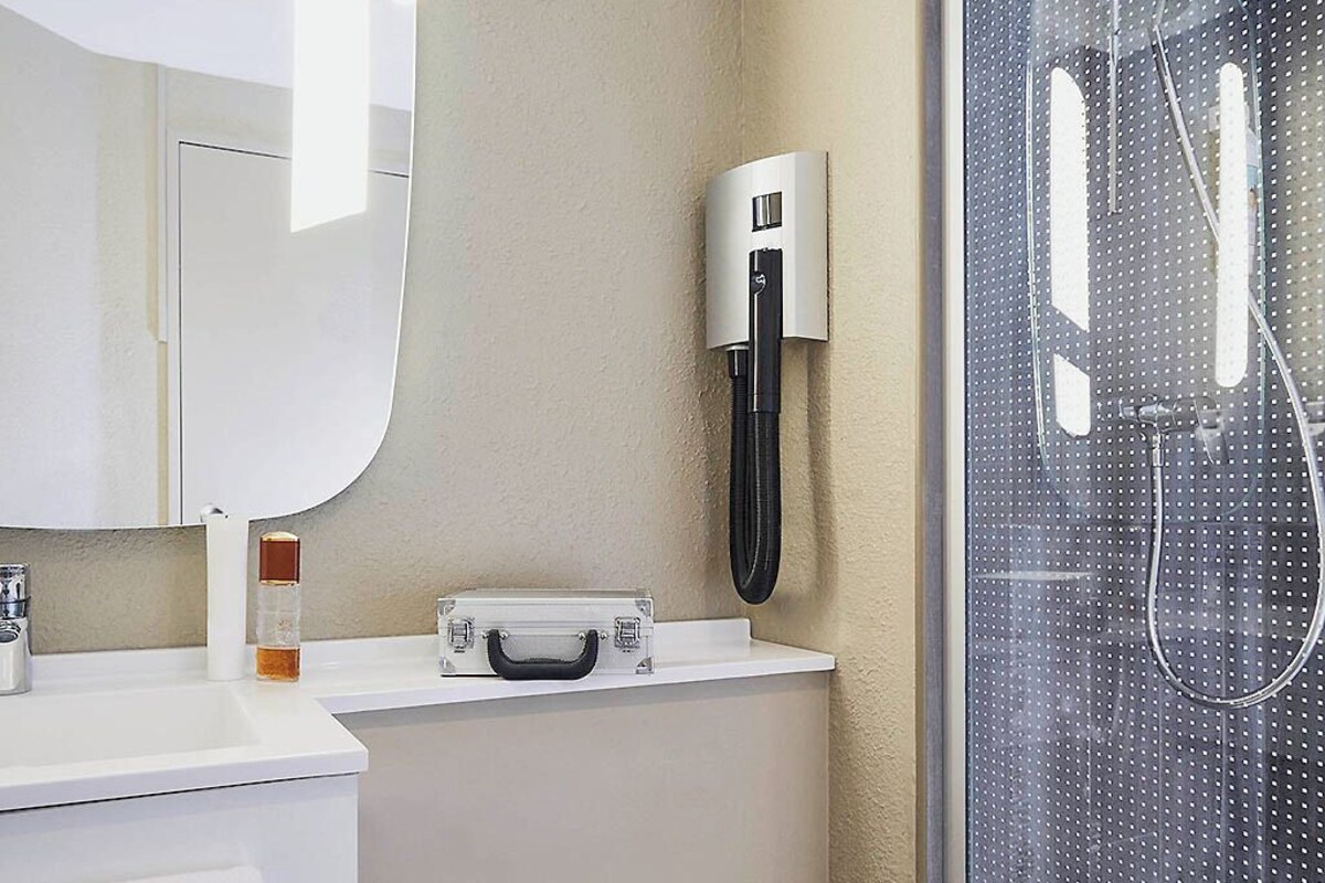 A bathroom with a white sink, a mirror, a towel dispenser, a shower with a glass door, and a wall-mounted faucet.