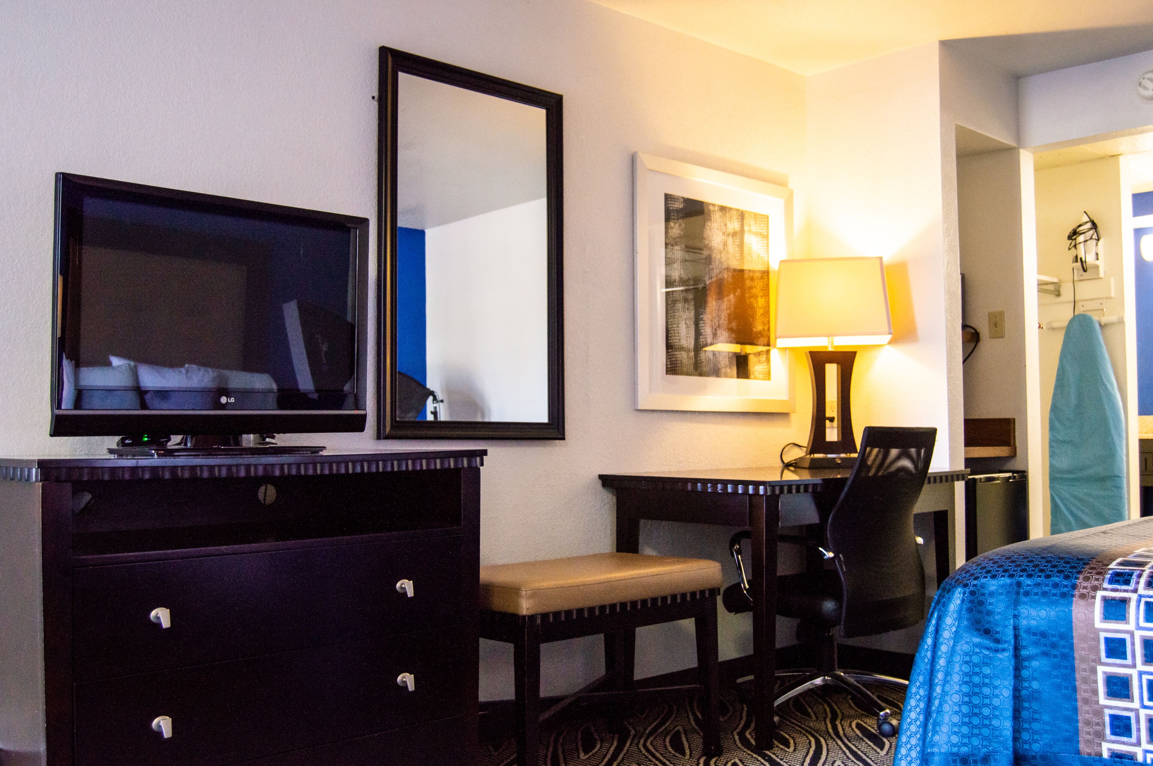 Habitación de hotel con televisión de pantalla plana, escritorio con silla, lámpara y cama con colcha de diseño azul y blanco.