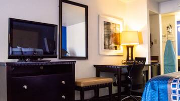 Habitación de hotel con televisión de pantalla plana, escritorio con silla, lámpara y cama con colcha de diseño azul y blanco.