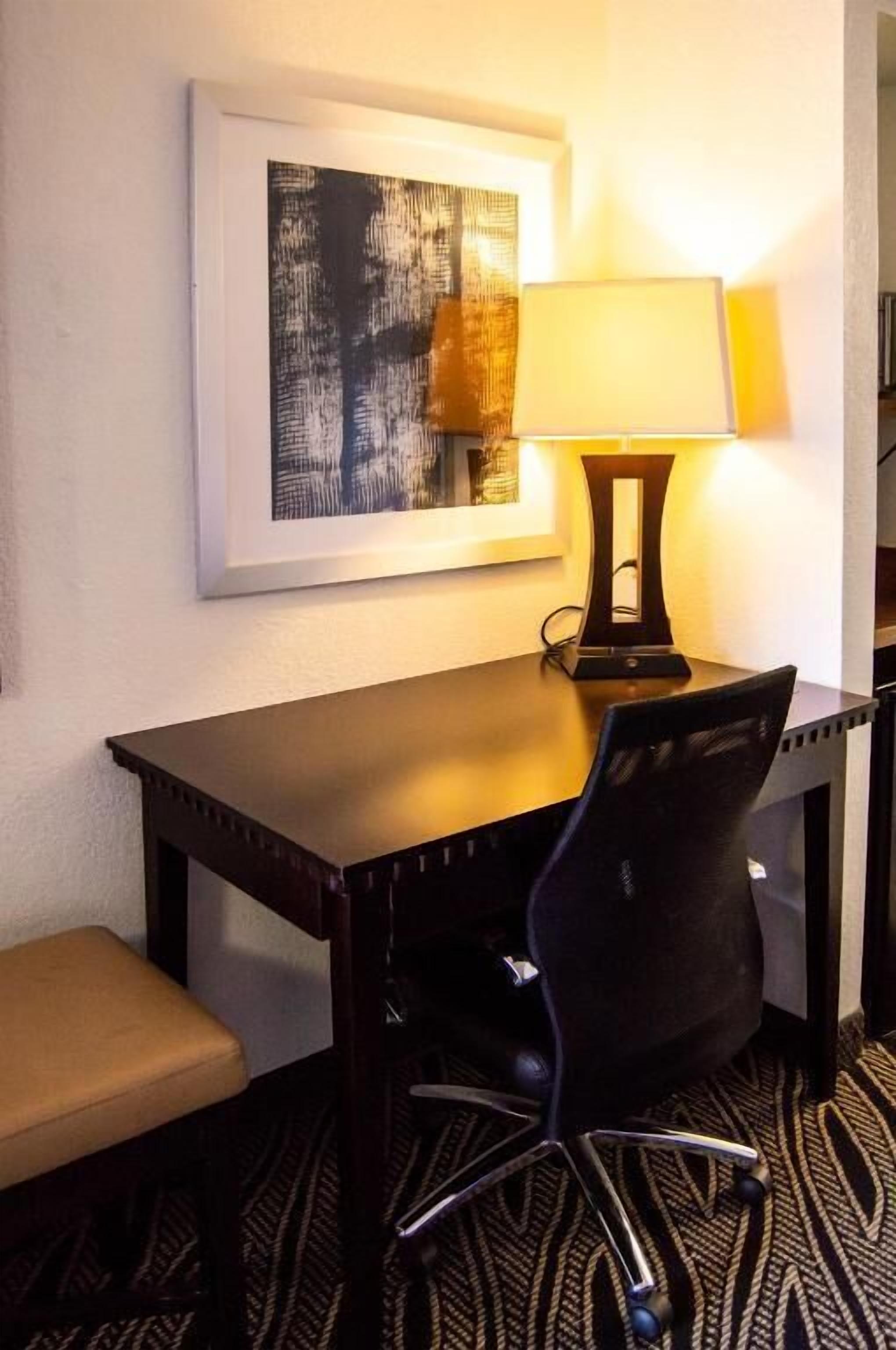 A hotel room with a desk, chair, and a framed abstract painting on the wall.