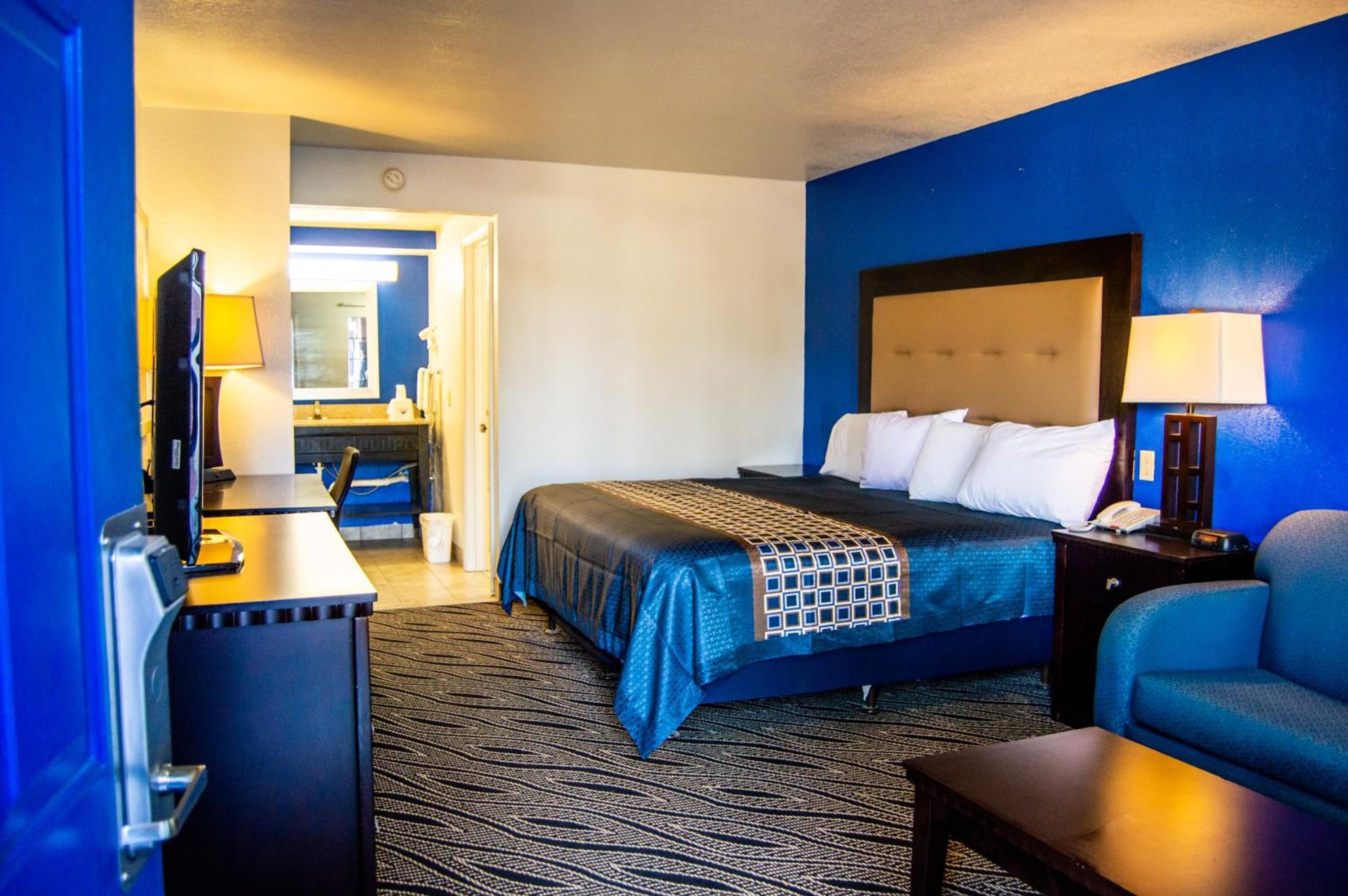 A hotel room with a bed, a desk with a computer, a chair, and a nightstand with a lamp.