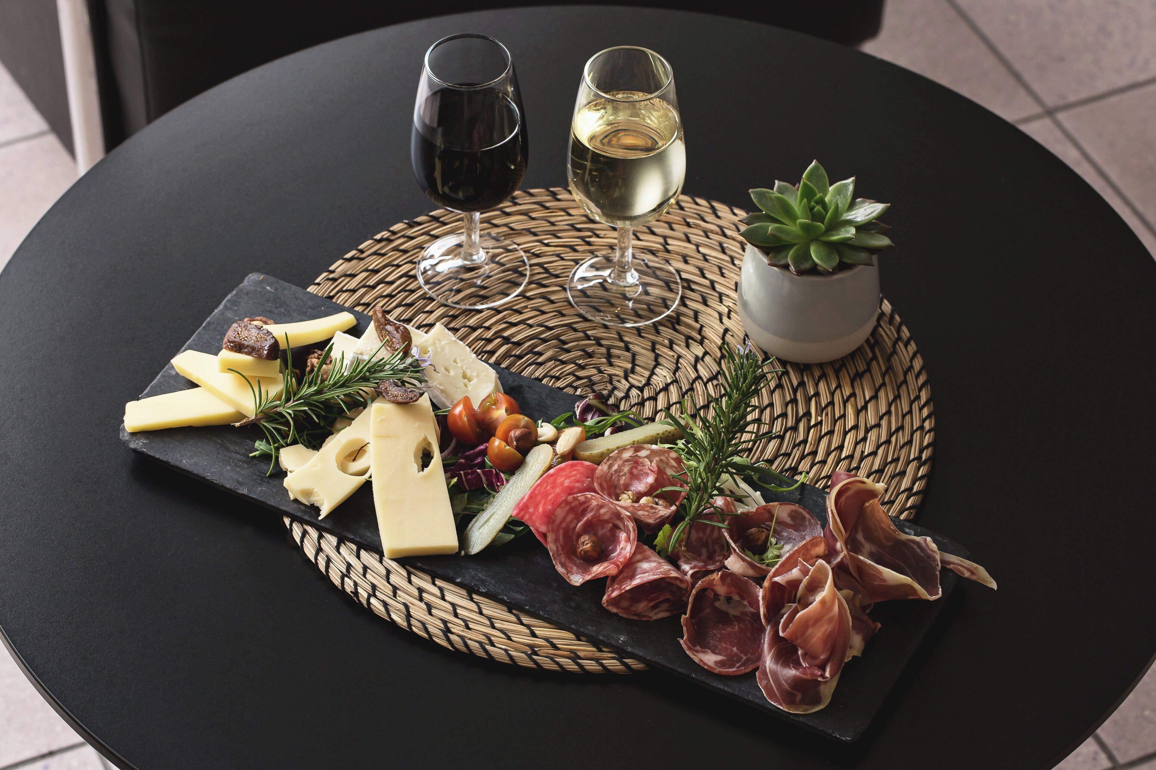 Una mesa redonda con una tabla de pizarra negra que sostiene varios quesos, embutidos y una pequeña planta en maceta. También hay dos copas de vino blanco sobre la mesa.